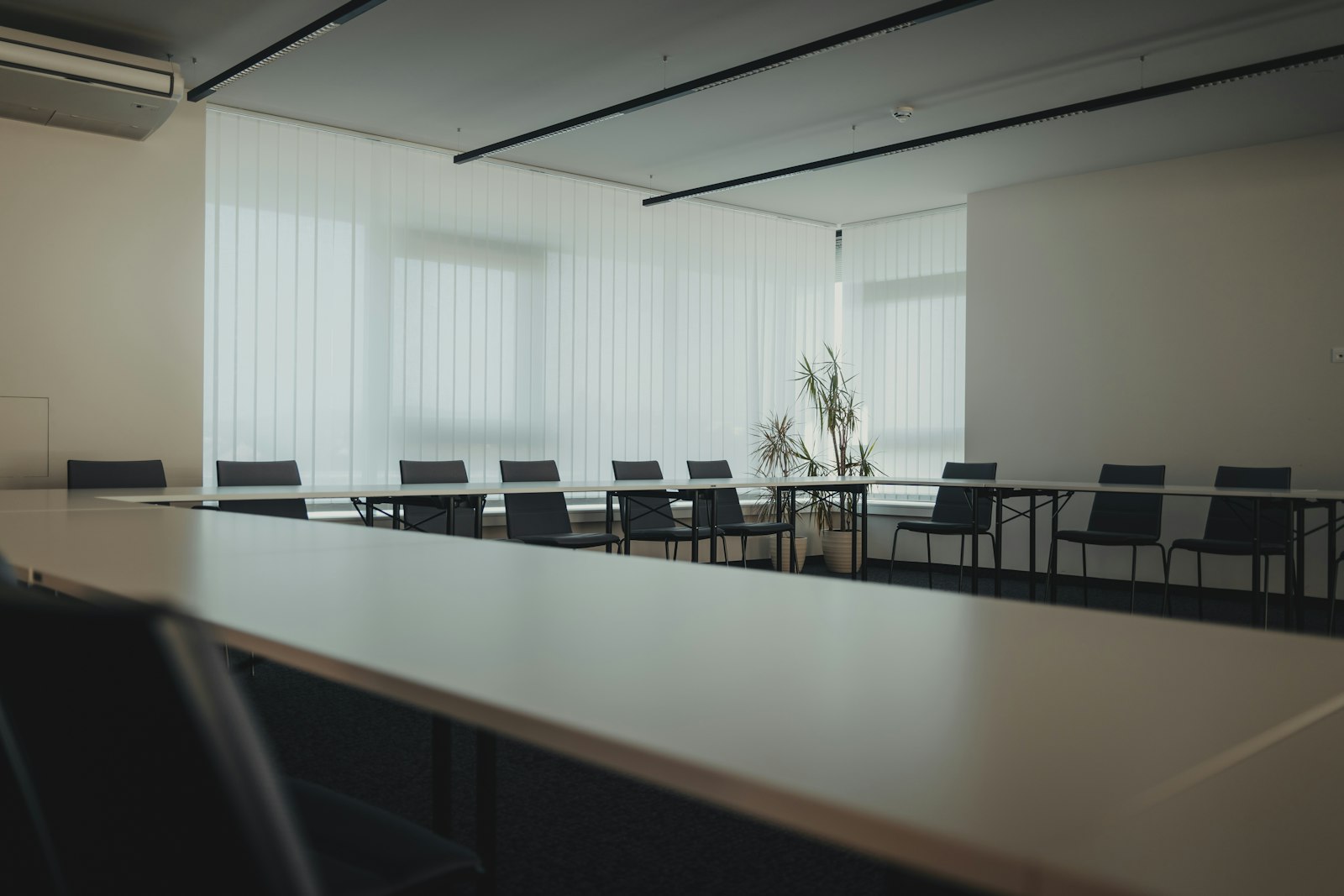 An empty meeting room with long white tables arranged in a U shape, black chairs and pale vertical blinds filtering daylight
