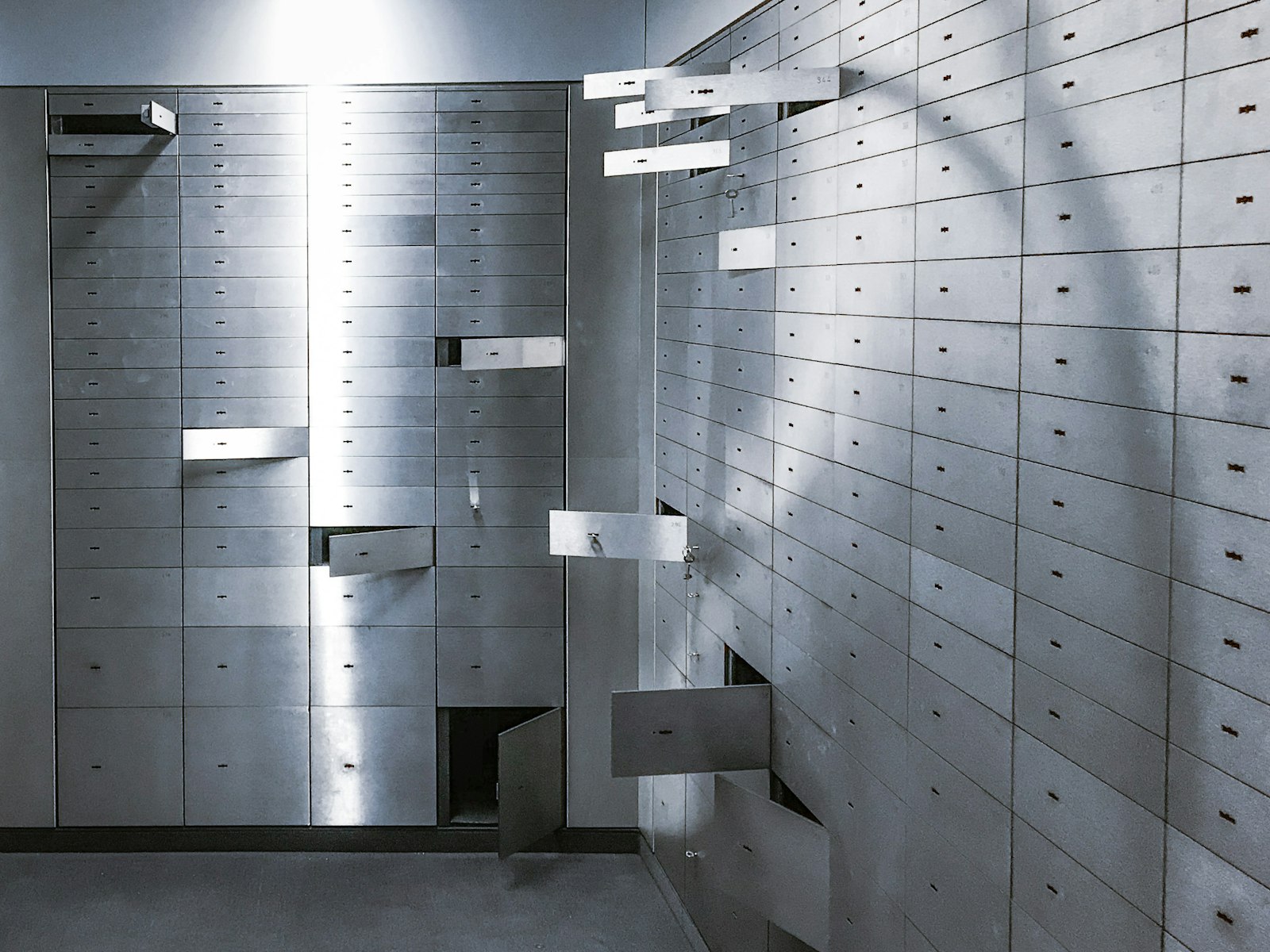 Rows of steel safe deposit boxes lining the walls of a bank vault, with several small doors standing open and a band of light falling across them