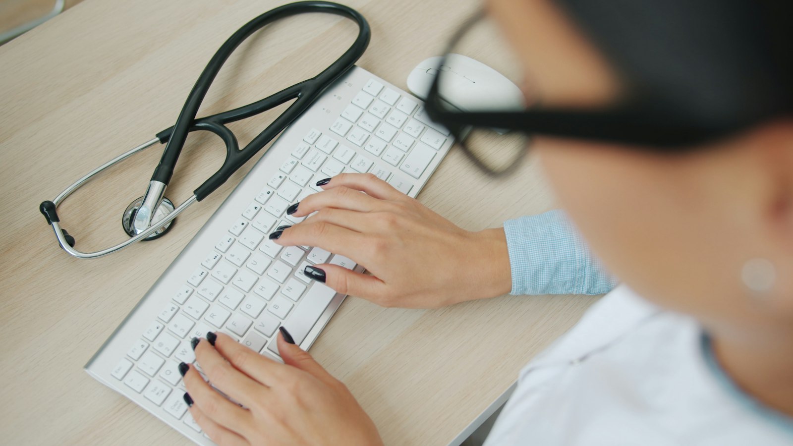 A doctor in glasses types on a white keyboard at a light wooden desk, seen from above, with a stethoscope lying beside the keyboard