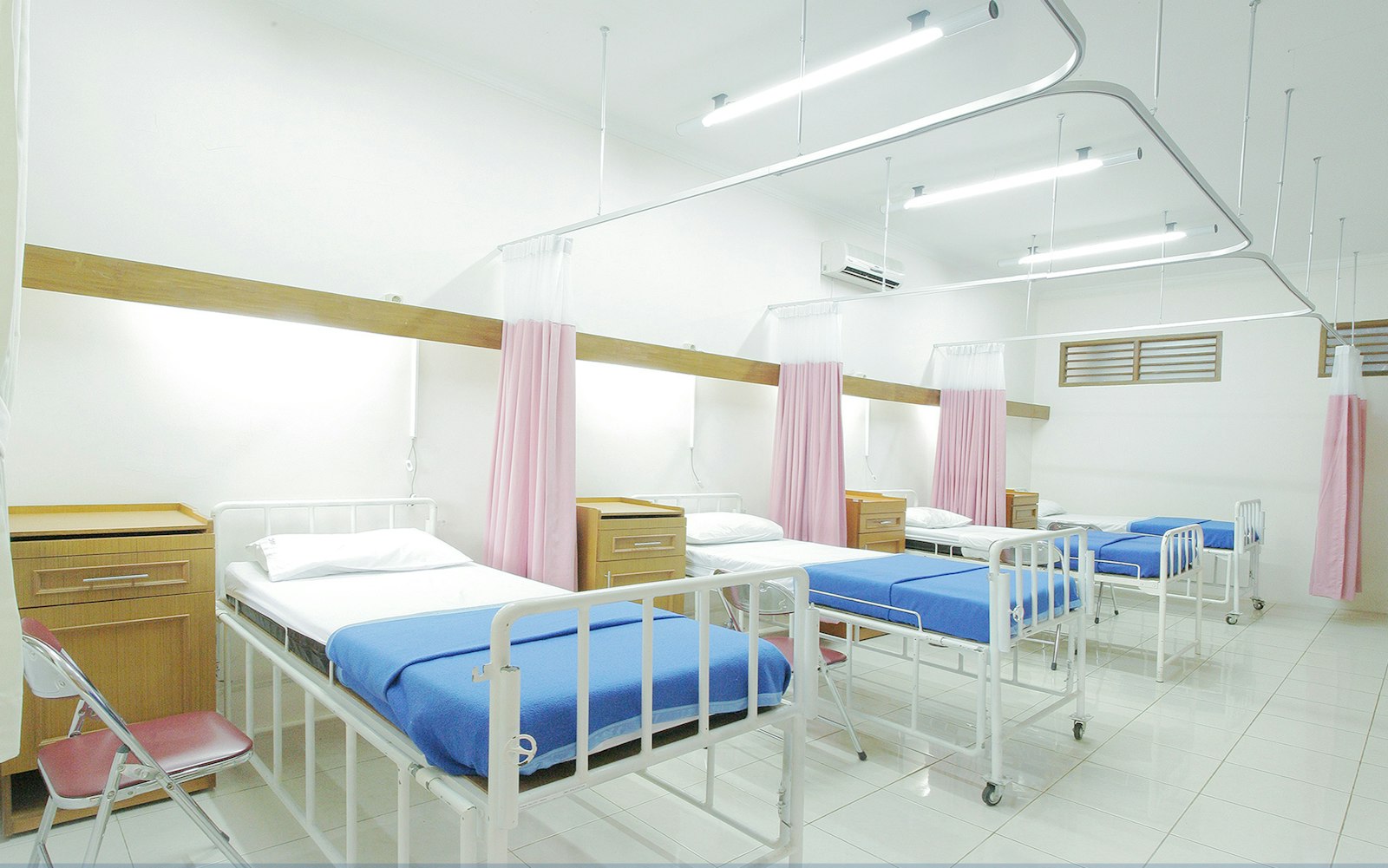 An empty hospital ward with made beds, blue blankets, wooden bedside cabinets and pink privacy curtains hanging from ceiling rails