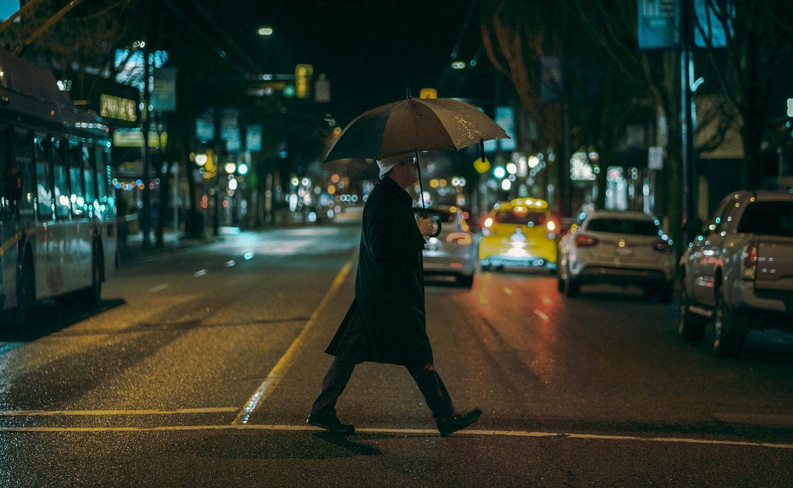 A person in a dark coat holding an umbrella crosses a wet city street at night, with a bus, a taxi and parked cars lit by streetlights behind them