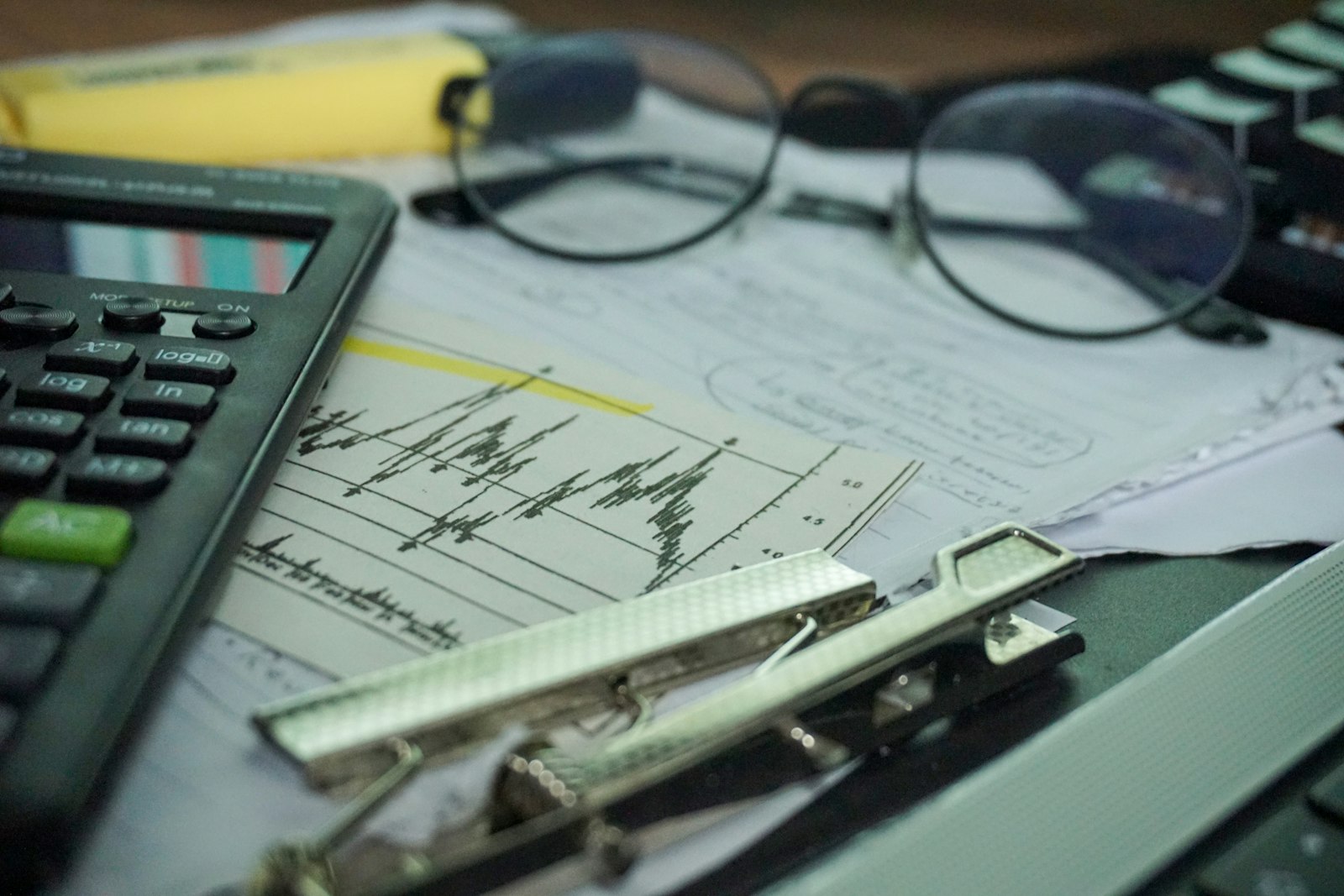 A close-up of a desk with a printed line chart, a pair of round glasses, a scientific calculator, a highlighter and a metal clipboard clip resting on the papers