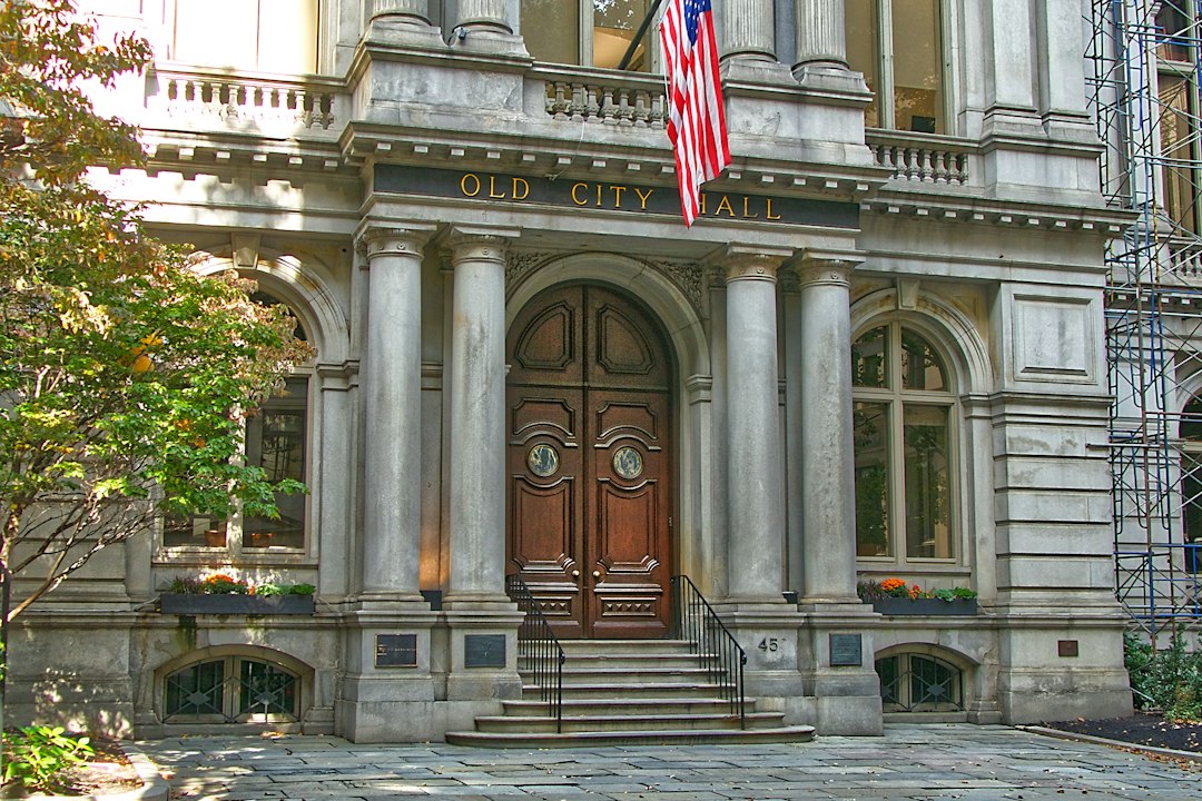 The arched wooden doors and stone facade of an old city hall with an American flag above the entrance