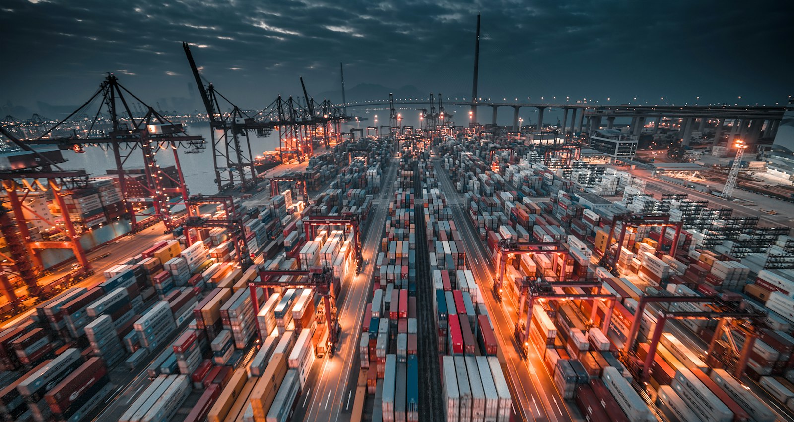 A container terminal at dusk seen from above, with stacked shipping containers in long rows, lit gantry cranes along the quay and a bridge in the background