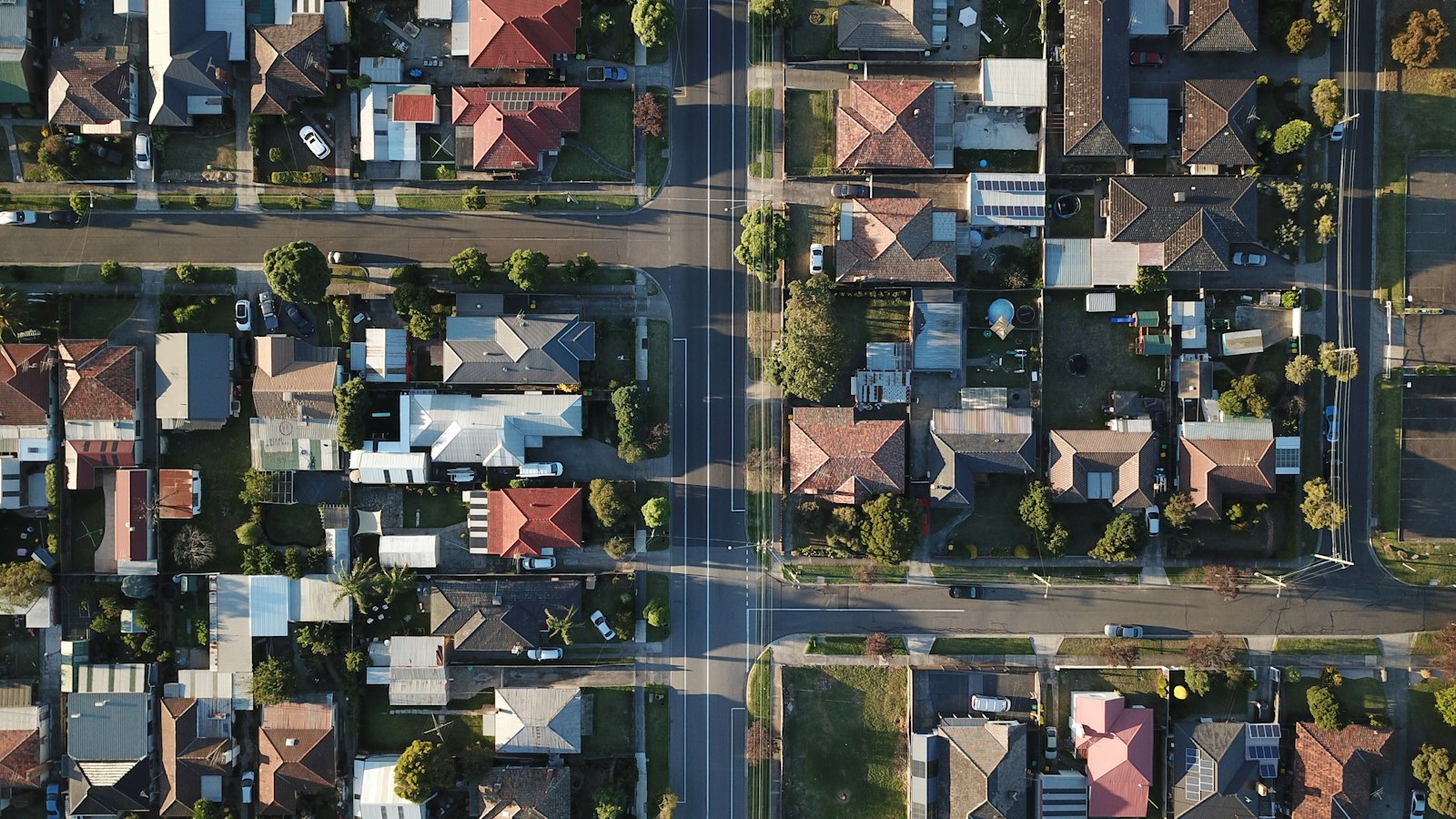 Aerial view straight down onto a suburban neighbourhood of detached houses with tiled roofs, gardens and parked cars on either side of a long straight road