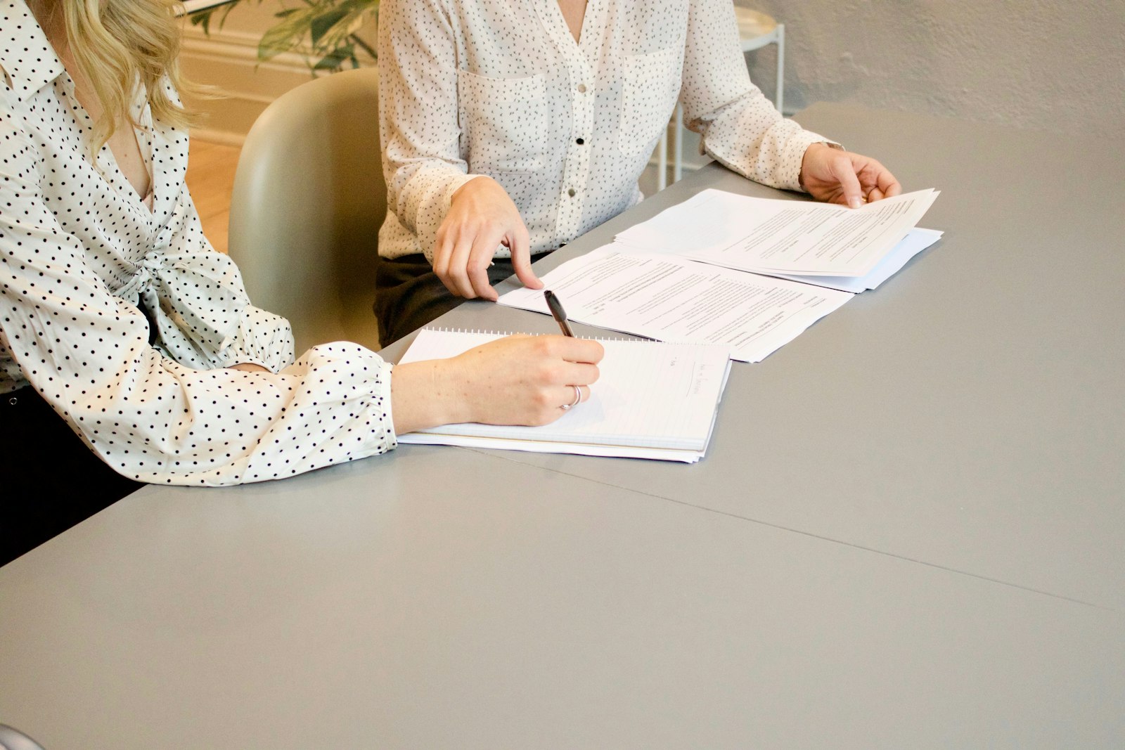 Two women in polka-dot blouses at a grey table, one writing in a notebook with a pen while the other points at a printed document