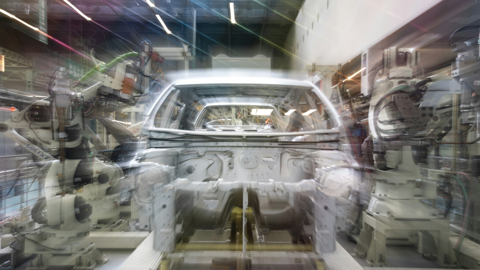 A bare car body shell on an assembly line between white industrial robot arms, photographed with a long exposure so the moving robots appear blurred