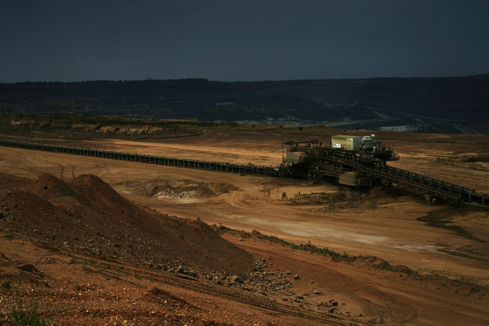 A long conveyor belt and a spreader machine crossing the reddish-brown floor of an open-pit mine under a dark, overcast sky