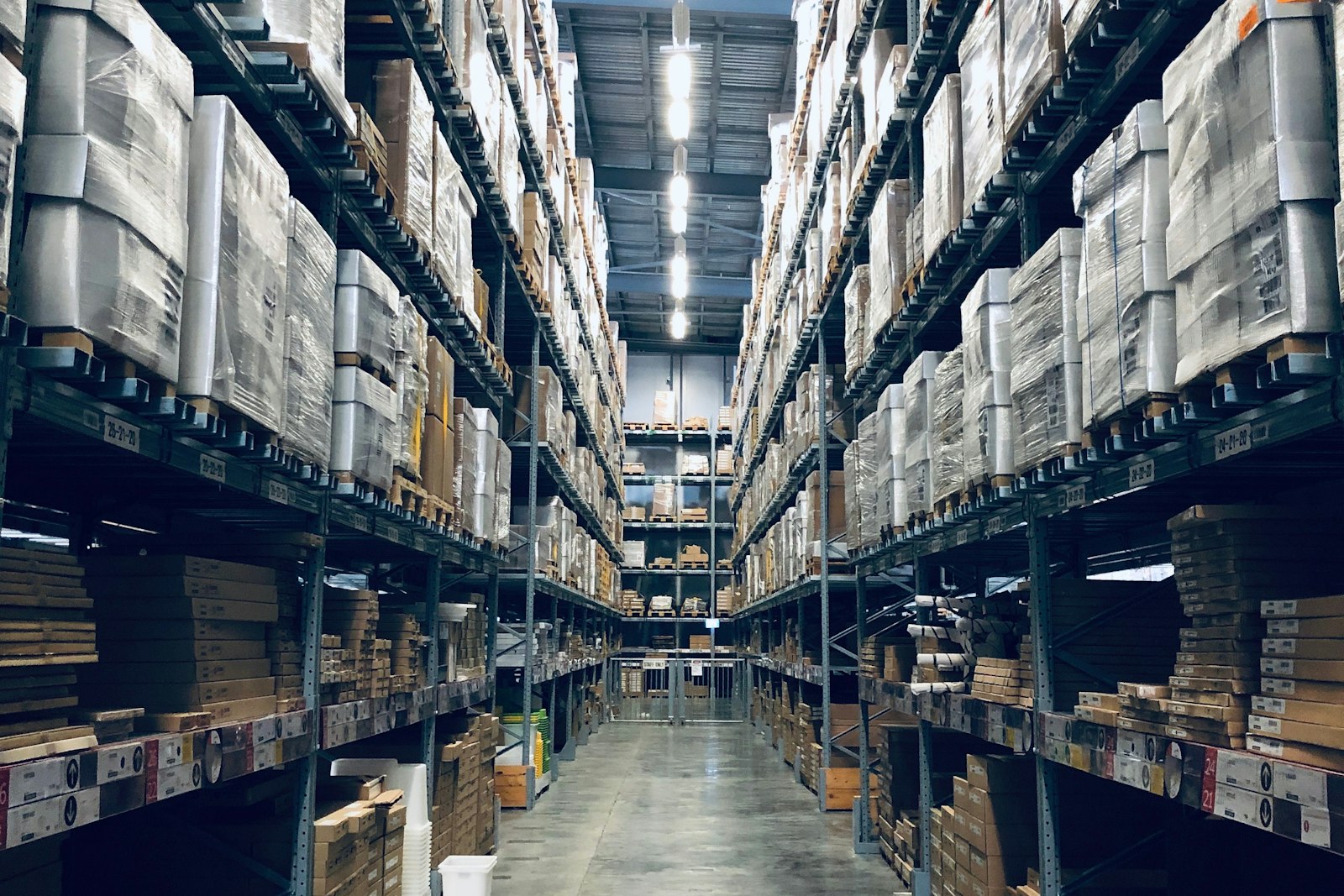 A long warehouse aisle between tall steel racks stacked with shrink-wrapped pallets and flat-packed cardboard boxes under bright ceiling lights