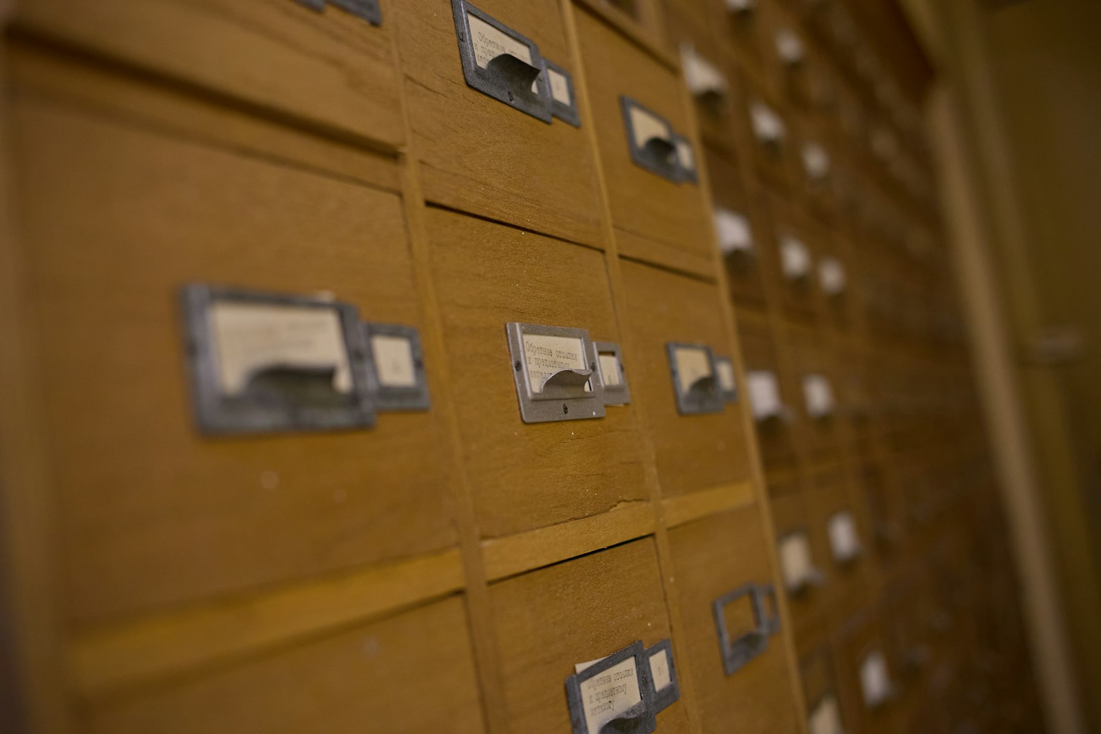 A wall of wooden library card catalogue drawers with metal label holders, receding out of focus into a dim corridor
