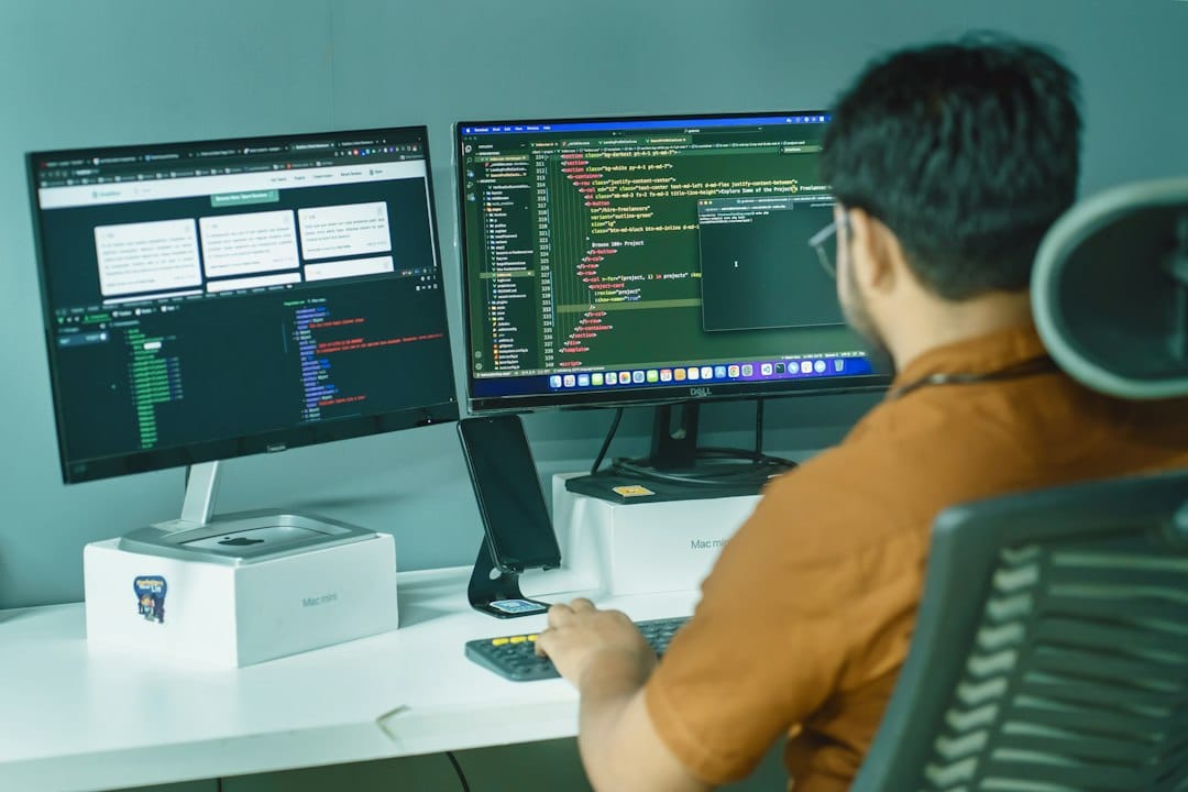 An engineer at a desk working across two monitors, one showing a terminal and one showing code, the kind of setup where adversarial test runs against an AI application are launched and reviewed