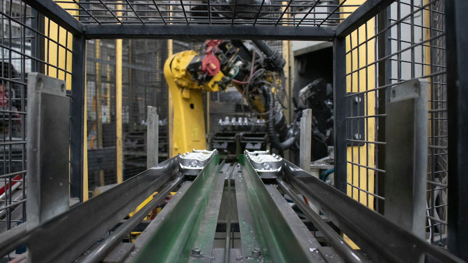 View along a factory conveyor with two green rails carrying metal parts towards a yellow industrial robot arm inside a wire safety cage