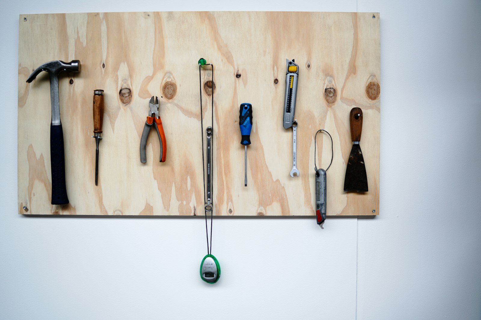 A row of hand tools, including a hammer, pliers, screwdriver, spanner, utility knife and scraper, hung neatly on a plywood board against a white wall