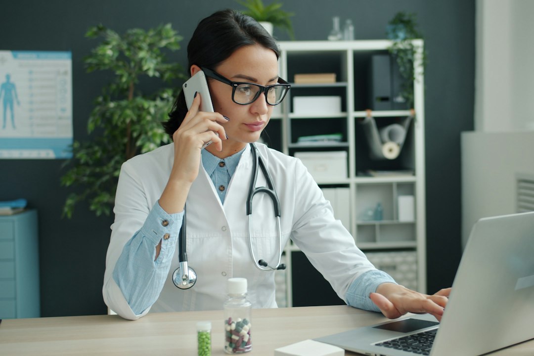 A physician in a white coat takes a phone call while typing on a laptop in a clinic office, where a locally hosted medical language model could draft notes and letters