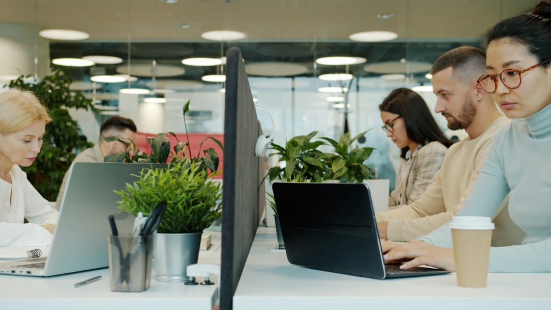 Five colleagues working on laptops at shared desks with plants and screen dividers in a bright open-plan office, the kind of staff an internal ChatGPT-style assistant is rolled out to