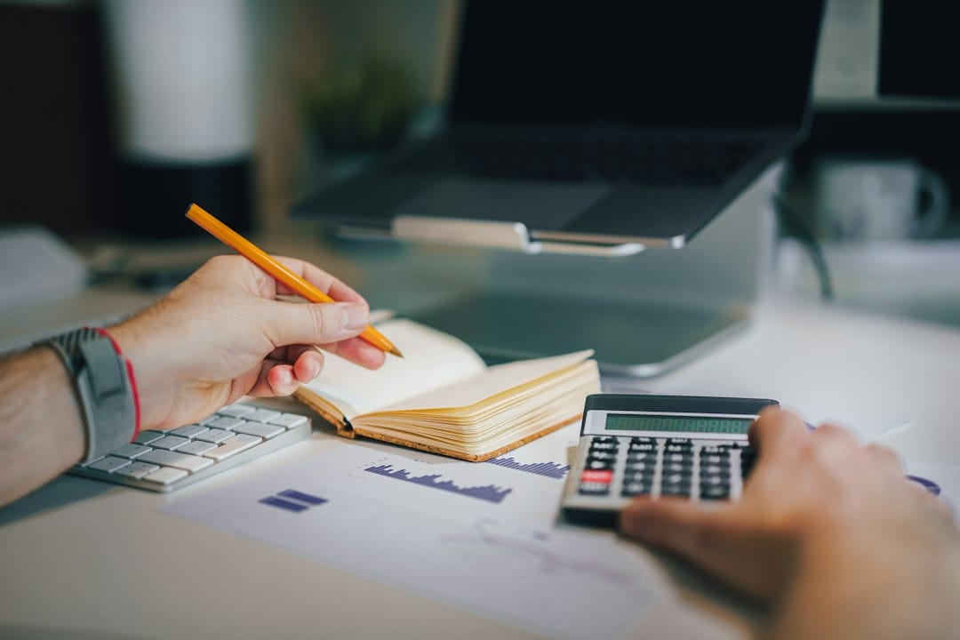 A person holding a pencil over an open notebook while working a calculator beside a laptop stand and printed bar charts, like a buyer totting up a year of ChatGPT and Copilot seats