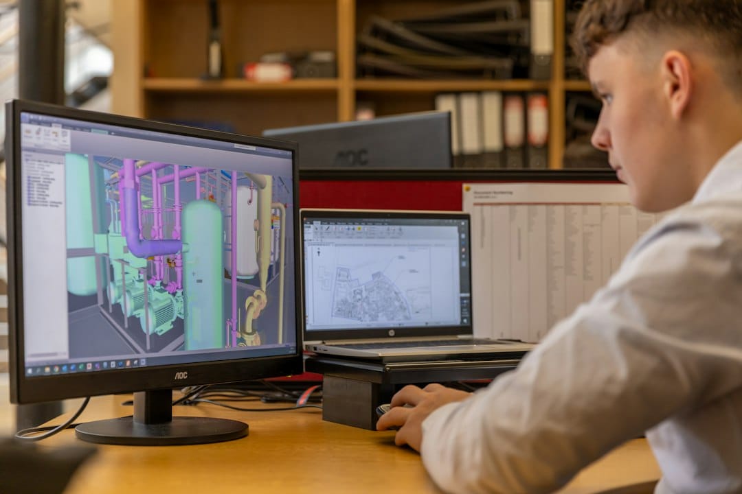 An engineer at a desk studies a 3D CAD model of plant piping on one monitor and a site drawing on a laptop, the kind of technical data defense contractors must keep inside a CMMC-assessed environment when they use AI tools