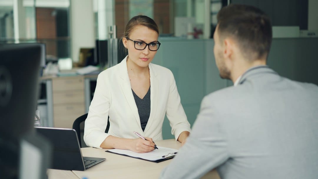 A recruiter in glasses takes notes on a clipboard beside her laptop while interviewing a candidate across an office desk, the kind of hiring step the EU AI Act treats as high-risk once AI scores or filters applicants