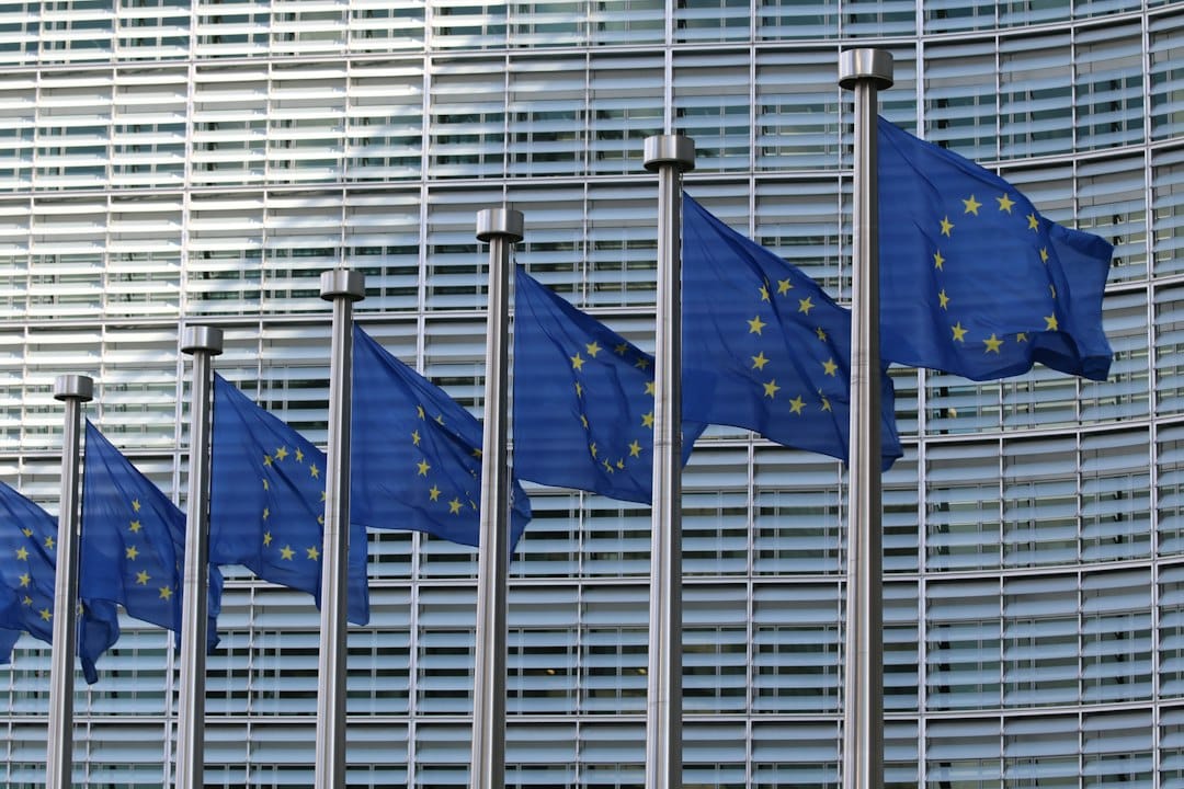 A row of European Union flags on tall poles in front of a large glass office building, used here to represent the EU data protection rules that decide which AI tools an organisation can use