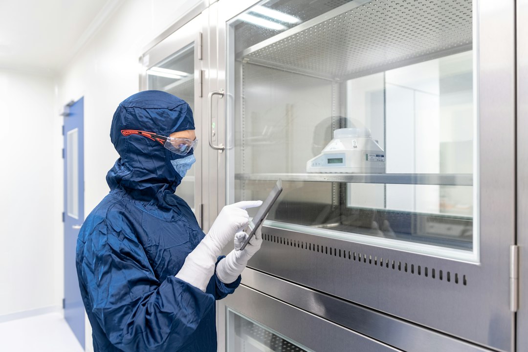 A cleanroom worker in a blue gown, mask and gloves checks a tablet beside stainless steel equipment in a pharmaceutical facility