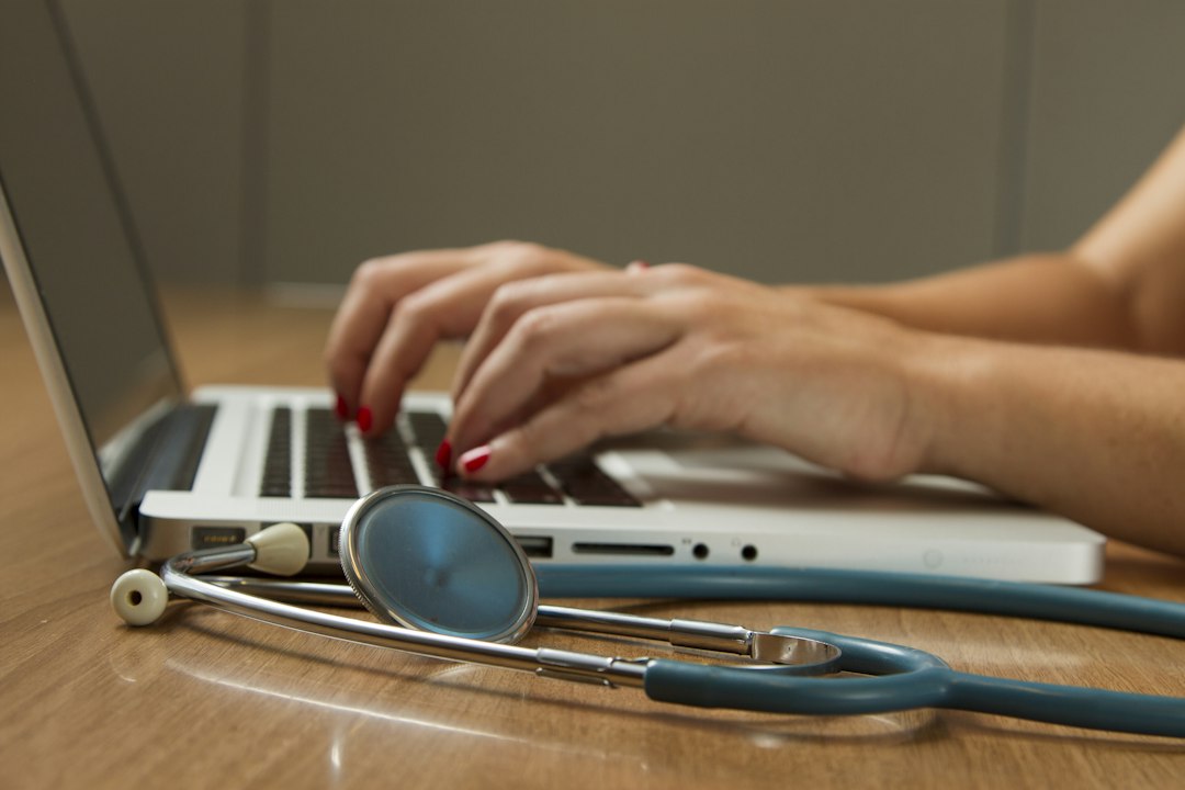 Hands typing on a laptop beside a stethoscope on a wooden desk, a reminder that prompts containing patient details become protected health information