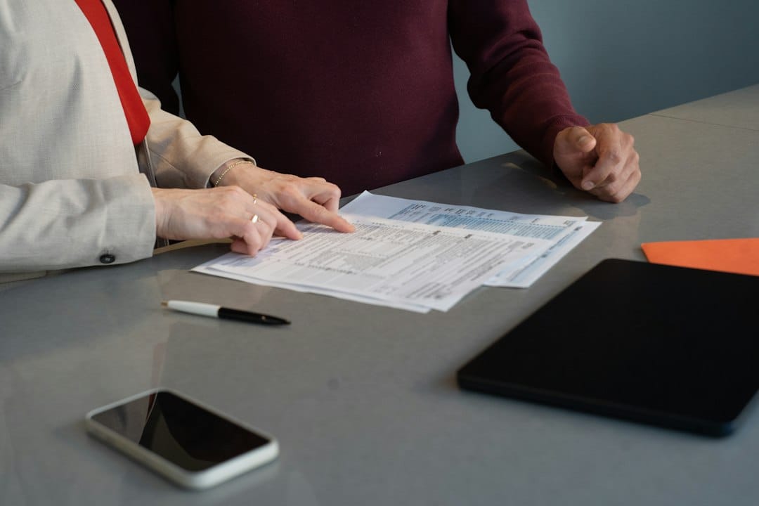 Two people at a grey table going over printed forms, with a smartphone and a tablet lying beside the papers, the kind of business information staff weigh up before pasting it into ChatGPT