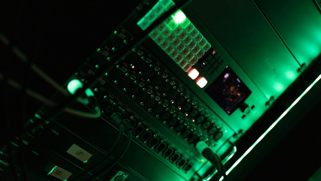 Rack-mounted equipment with rows of cable sockets, a lit button panel and a small screen glowing green in a dark equipment room, standing in for the in-house hardware a self-hosted Llama deployment runs on