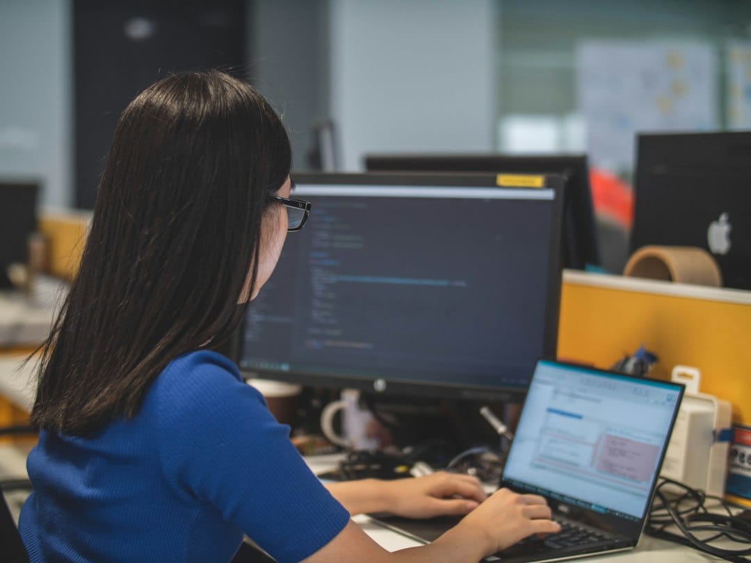 An engineer at an office desk working on a laptop in front of a larger monitor full of code, the kind of screen where LLM traces, token counts and latency charts get read