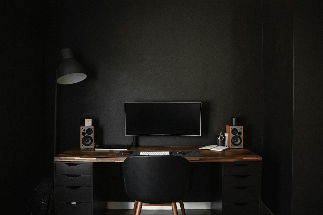A minimal home office in a dark room, with one monitor, a desk lamp and two small speakers on a wooden desk, the kind of setup where people run local AI agent apps