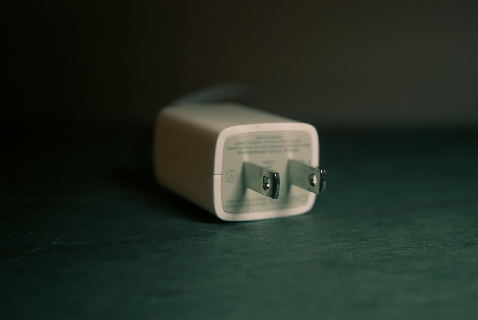 A small white two-pin power adapter lying on a dark green surface, photographed close up with a shallow depth of field