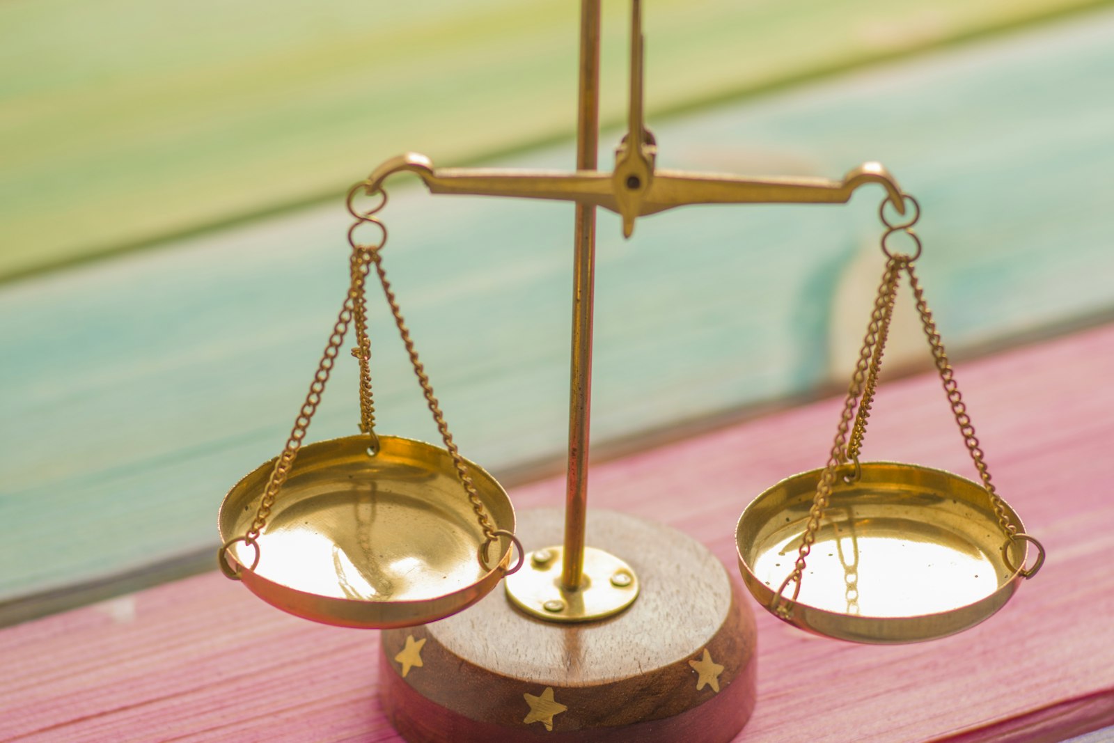 A small brass balance scale with two empty pans standing on a wooden base, set on painted boards in pink, teal and yellow