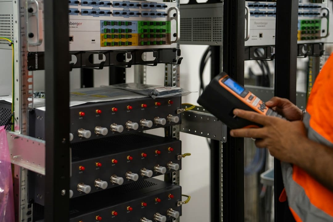 A technician in a high-visibility vest checks a rack of network equipment with a handheld test device, representing the network and information systems that NIS2 requires essential and important entities to secure
