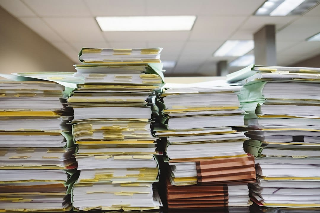 Tall stacks of paper files and folders flagged with yellow sticky notes on a desk under office ceiling lights, the kind of backlog an on-premise document AI pipeline is built to read