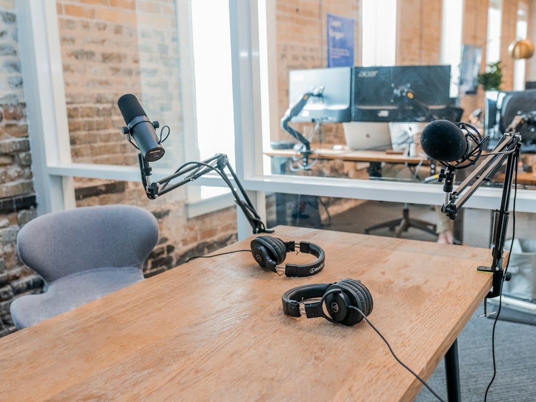 Two microphones on boom arms and two pairs of headphones on a wooden table in a glass-walled office room, standing in for the capture and playback ends of a voice AI system that stays inside the building