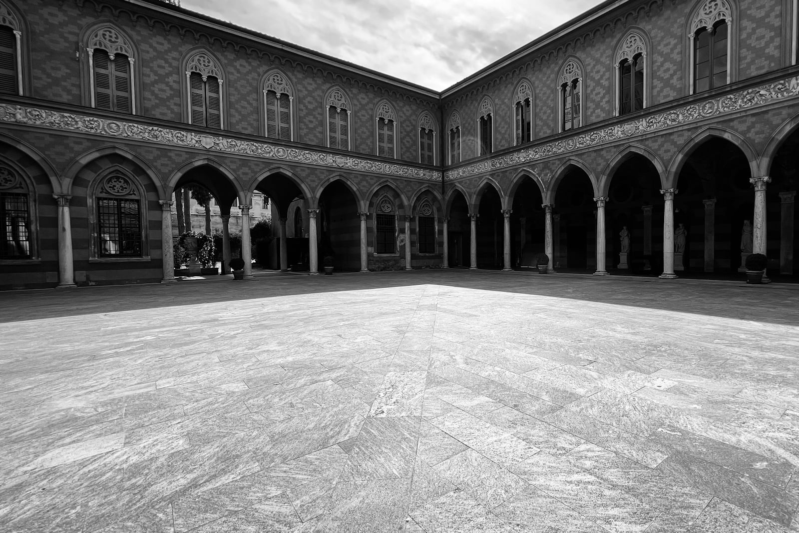 Black and white photo of an empty paved courtyard enclosed on two sides by arcaded buildings with pointed arches and shuttered windows, under a cloudy sky