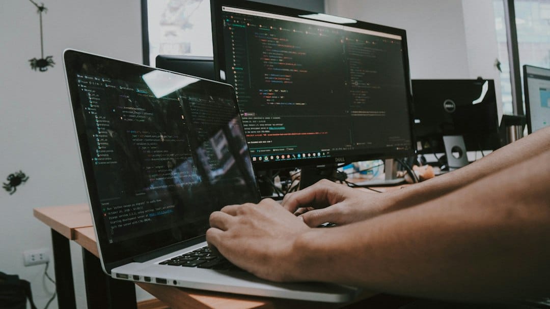 Hands typing on a laptop beside a desktop monitor, both screens showing code in a dark editor theme, the kind of workstation where engineers decide which tools an AI agent may call