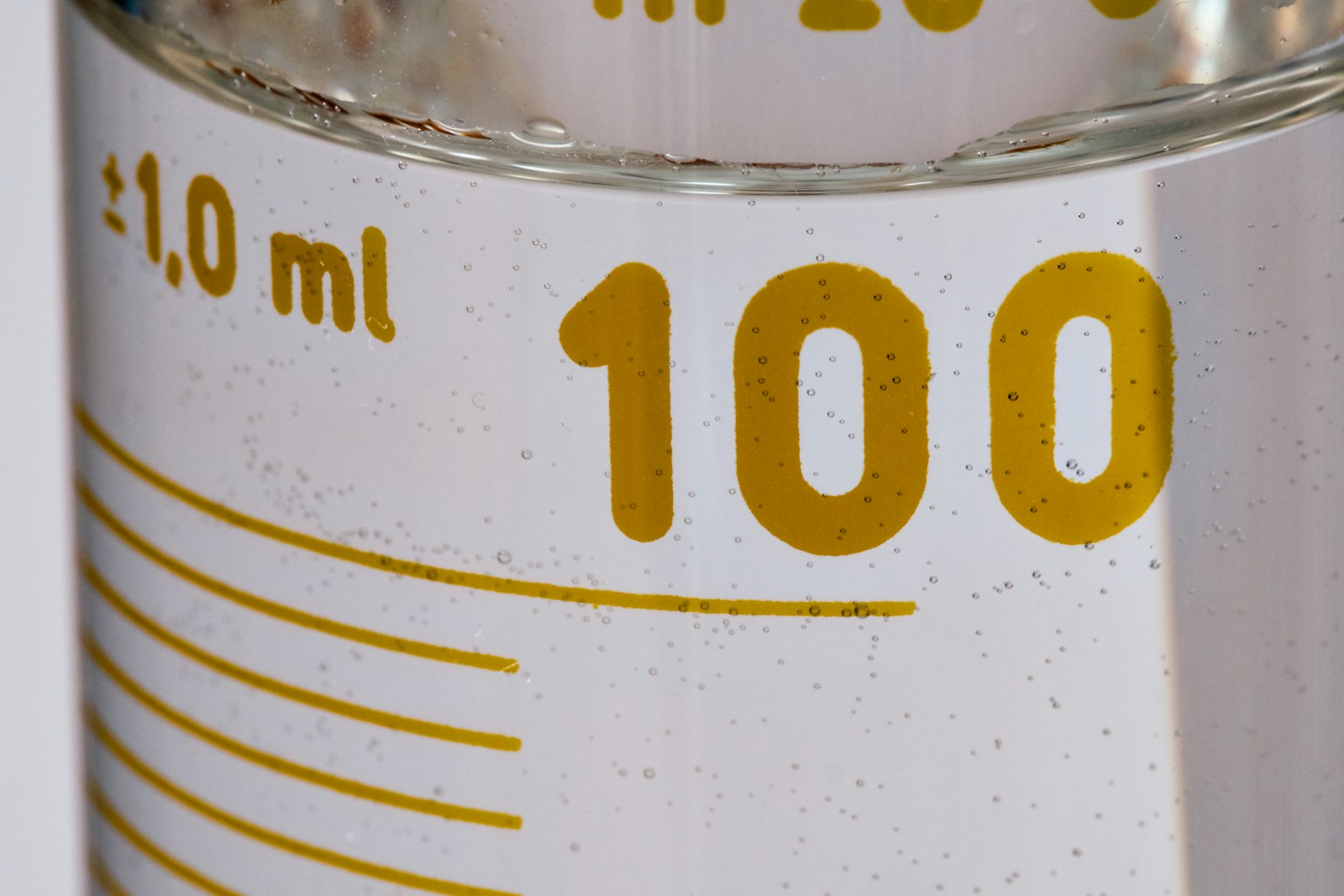 Close-up of a glass measuring cylinder filled with clear liquid and small bubbles, with yellow graduation marks and a printed tolerance of plus or minus one millilitre