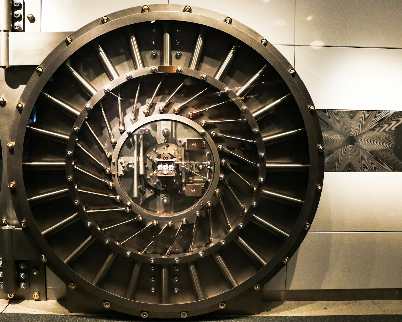 A round steel vault door set into a pale stone wall, with dozens of polished locking bolts radiating from a glass-covered locking mechanism at its centre