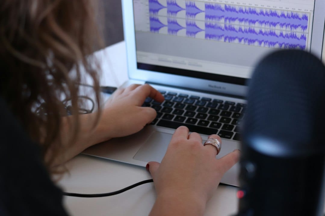 Over-the-shoulder view of a person typing on a laptop that shows purple audio waveforms in an editing program, with a desktop microphone in the foreground, like a recording about to be turned into a transcript